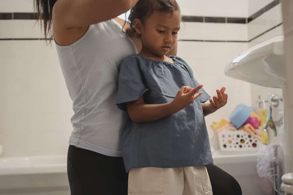 A mother carefully styles her young daughter's hair in a cozy, familiar bathroom setting.
