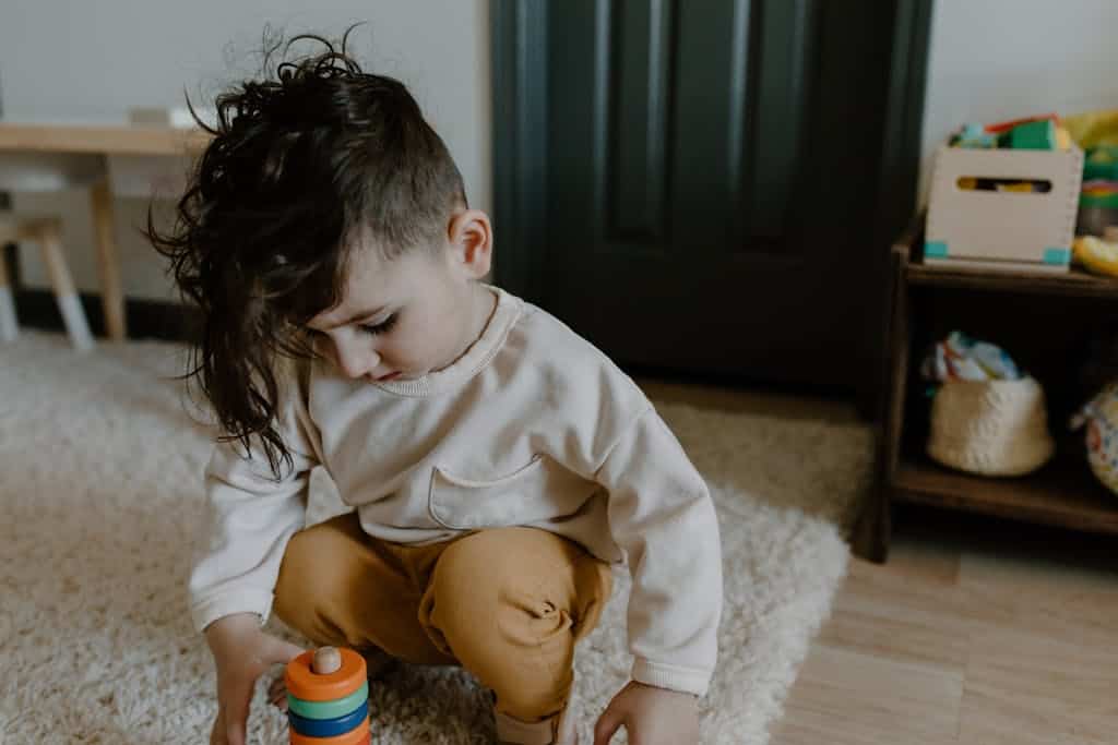 A young child with curly hair plays with a colorful stacking toy on a carpeted floor indoors.