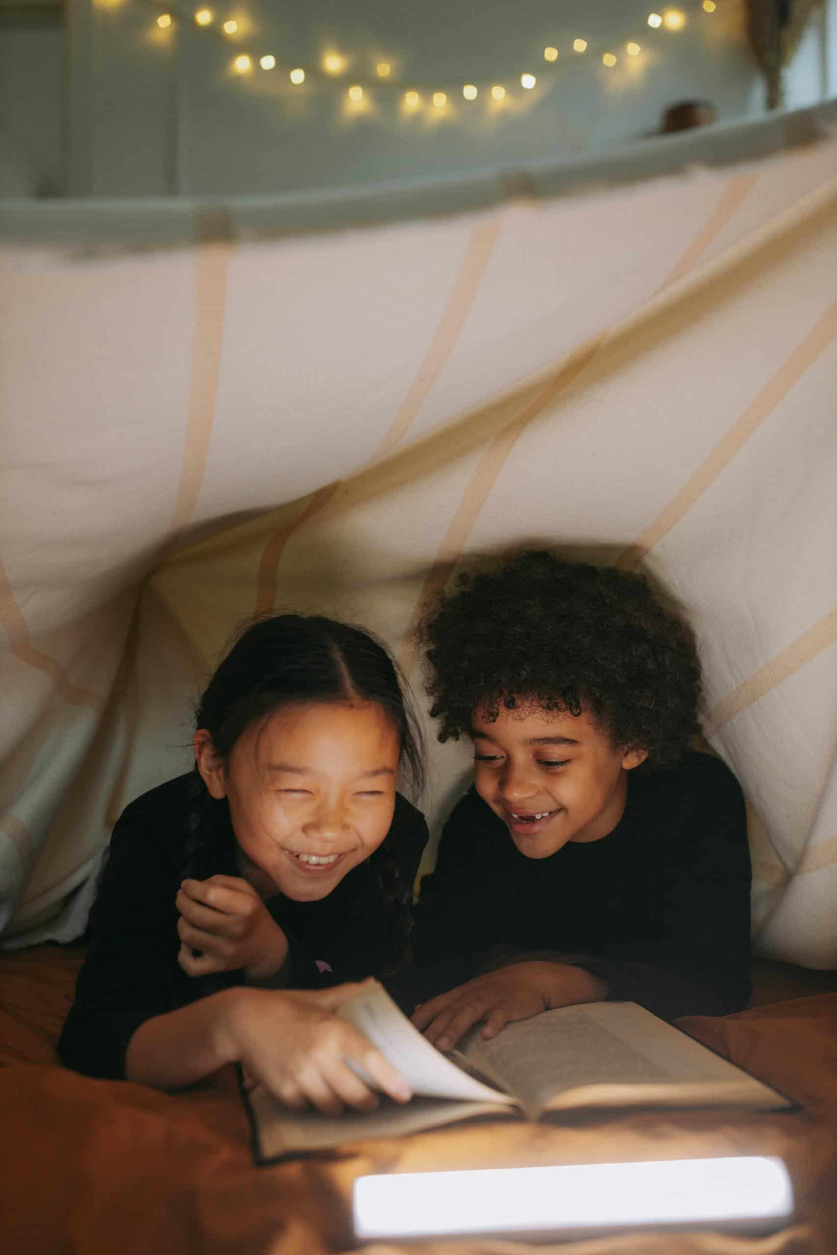 Two children happily reading under a cozy blanket fort with warm lighting.