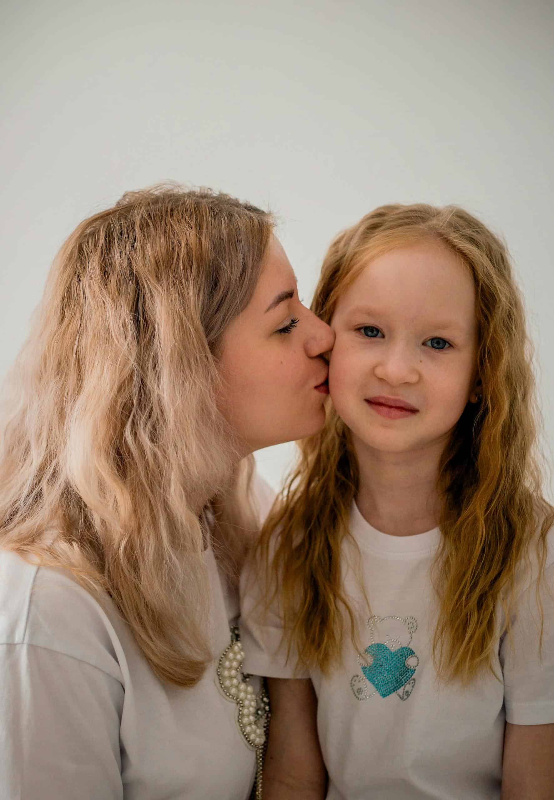 A mother lovingly kisses her daughter indoors, both wearing white shirts.