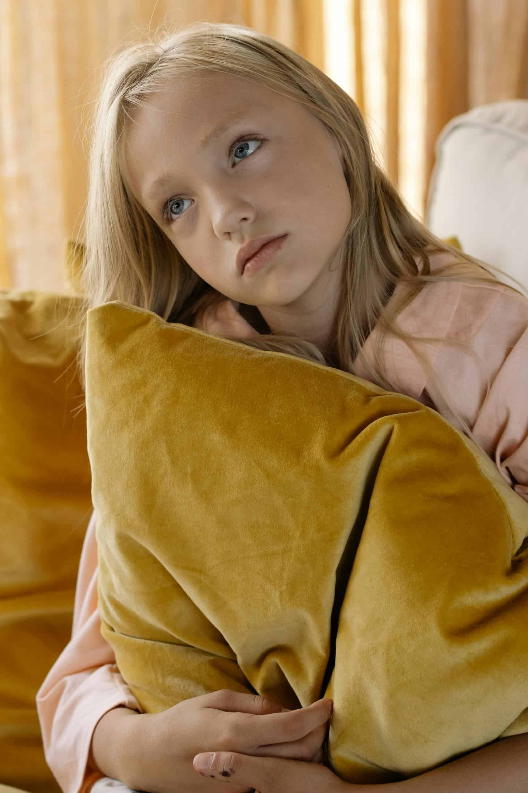 Pensive girl holding a yellow cushion, looking lost in thought indoors.